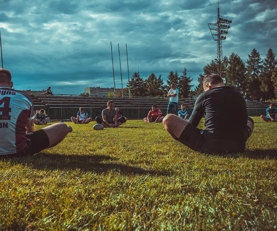 Fotografia eventowa prezentująca trening drużyny rugby. Zawodnicy siedzą na ziemii.
