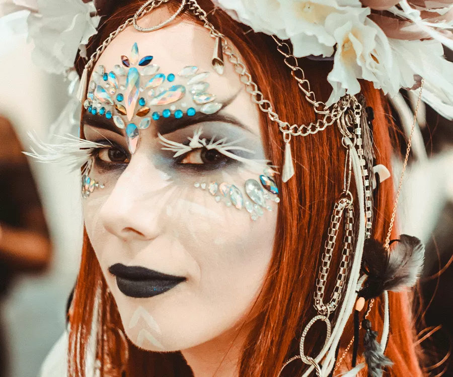 Fotografia eventowa prezentująca twarz dziewczyny w bogatym artystycznym makijażu z kryształami, piórkami i łańcuszkami