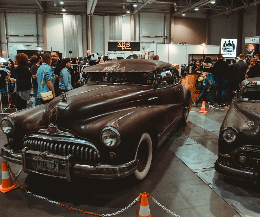 Fotografia eventowa prezentująca zabytkowe amerykańskie samochody w hali, w koło tłum ludzi. 