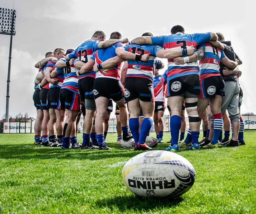 Fotografia eventowa prezentująca drużynę rugby w trakcie narady, na pierwszym planie znajduję się piłka do rugby.