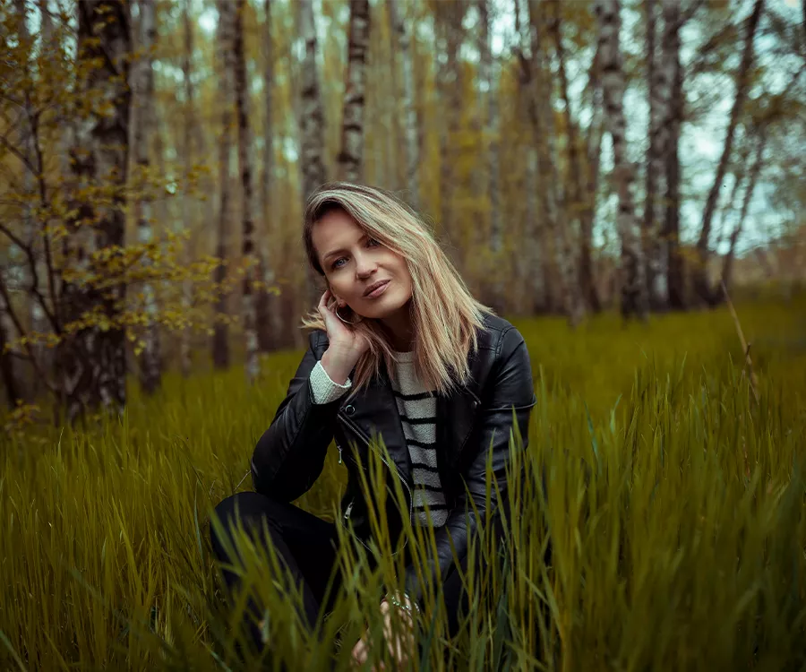 Blondynka siedząca w zielonej trawie, w tle las.