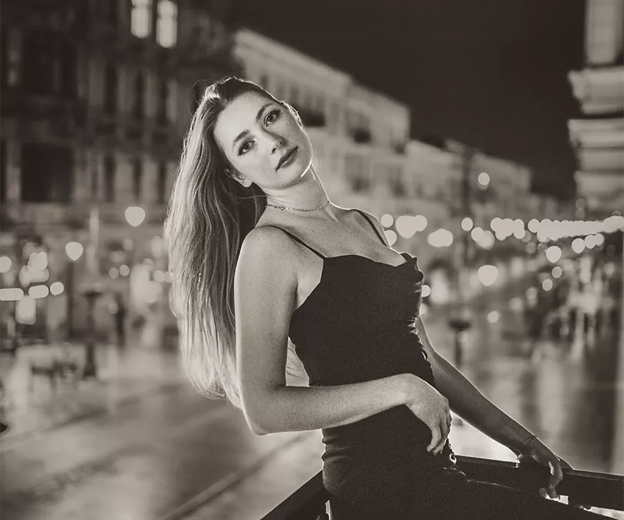 Nocna panorama miasta z kobietą w czarnej sukience, która elegancko pozuje na balkonie, tworząc tajemniczy klimat