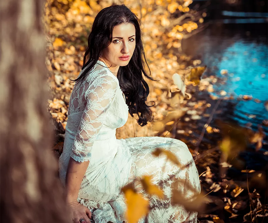 Piękna uśmiechnięta kobieta w koronkowej sukience siedzi na ziemi, otoczona delikatnym światłem i naturą.