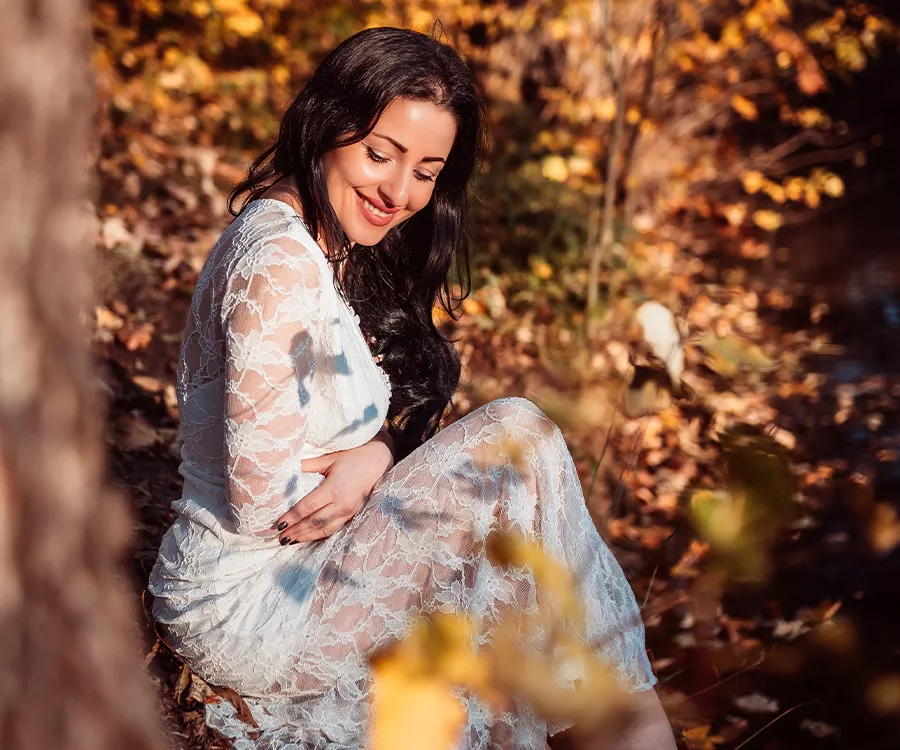 Piękna uśmiechnięta kobieta w koronkowej sukience siedzi na ziemi, otoczona delikatnym światłem i naturą.