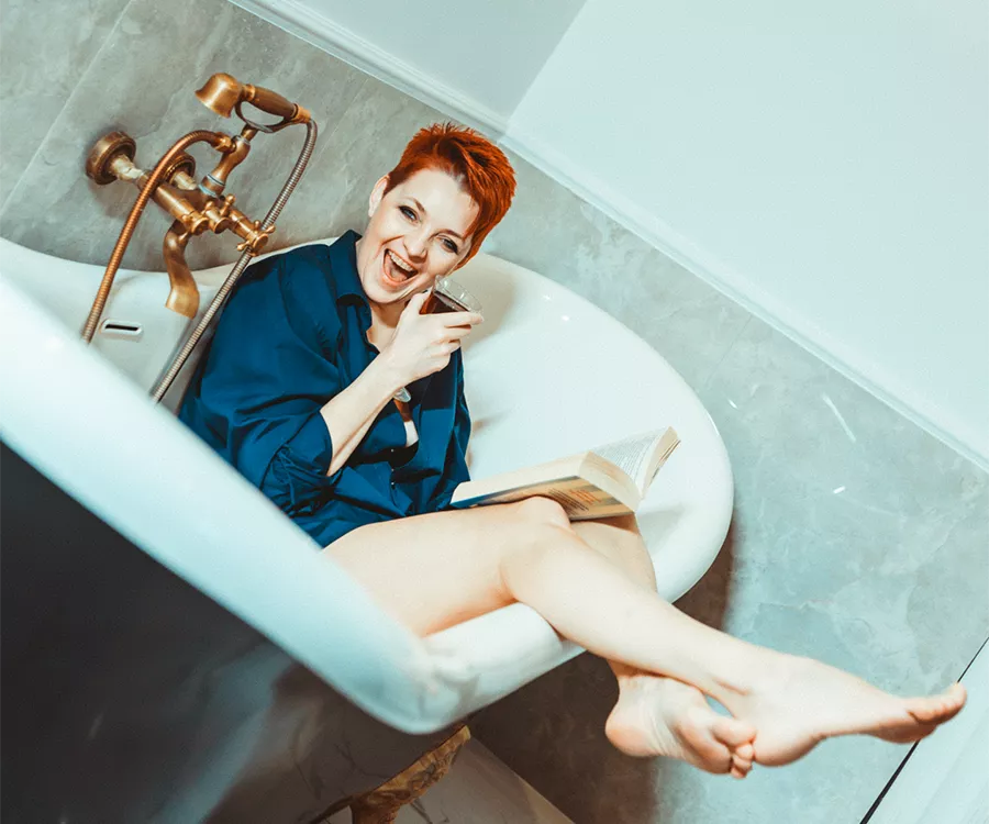 Roześmiana kobieta relaksuje się w wannie, trzymając książkę i kieliszek wina, ciesząc się chwilą spokoju.