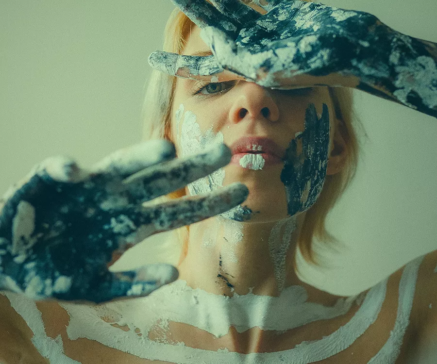 Portret dziewczyny, która trzyma dłonie przed twarzą. Jest umazana farbą.