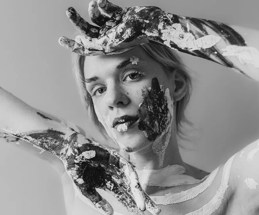 Czarnobiały portret dziewczyny, która trzyma dłoie przez twarzą. Jest umazana farbą.