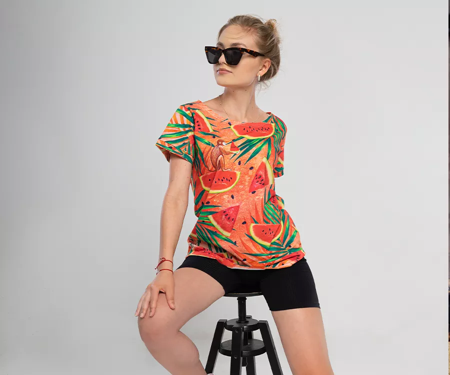 Zdjęcie produktowe przedstawiające dziewczynę siedzącą na stołku, w okularach przeciwłonecznych. Ubrana w koszulkę AOP w motywy owocowe