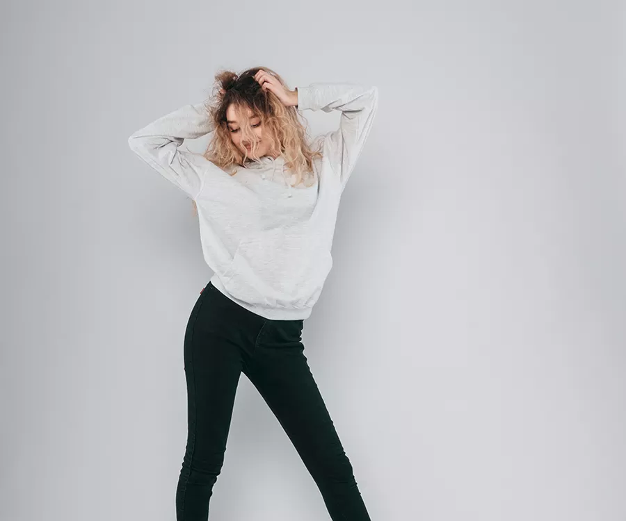 Zdjęcie produktowe przedstawiające dziewczynę w studiu fotograficznym, pozującą na białym tle. Ubrana w ciemne jeansy i jasną bluzę.