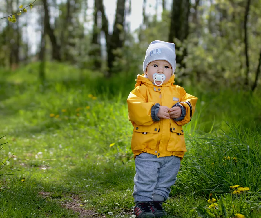 Zdjęcie przedstawiające małego chłopca ubranego w intensywnie żółtą kurtkę. Znajduję się on na leśnej ścieżce. 