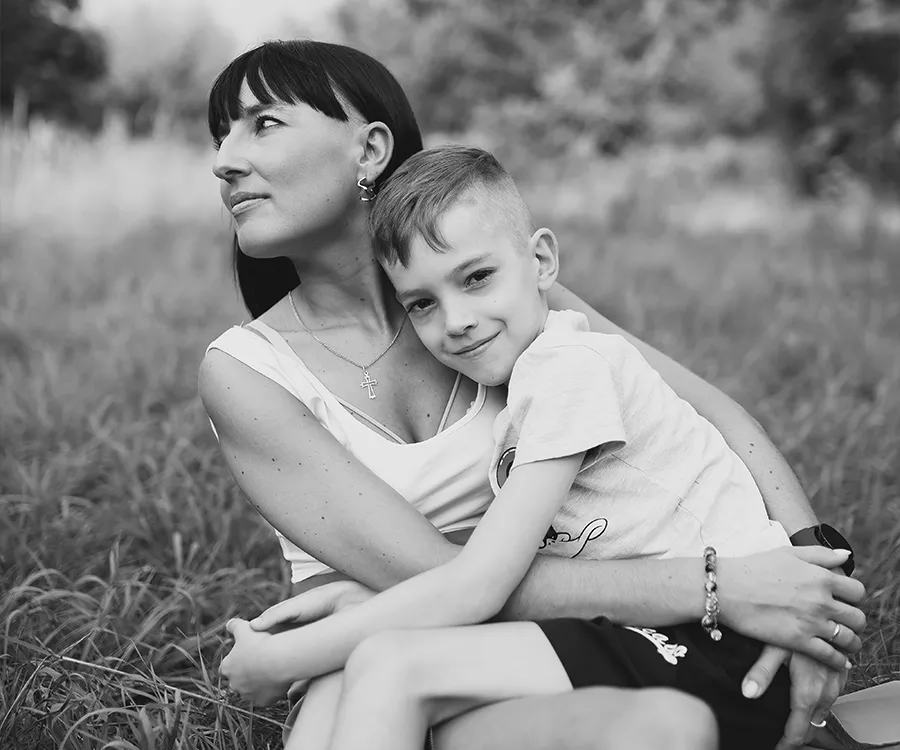 Fotografia mamy z synem na kolanach, siedzą na trawie pośród łąk. Zdjęcie w odcieniach szarości. 