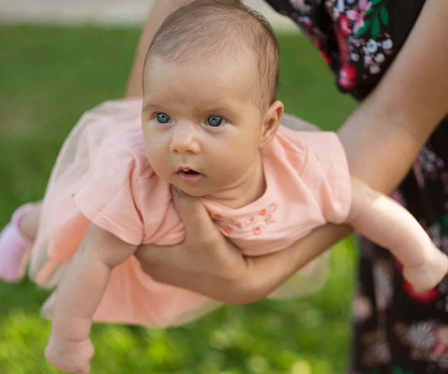 Zdjęcie kilku miesięcznej dziewczynki w różowej sukience, trzymanej na dłoniach przez mamę.