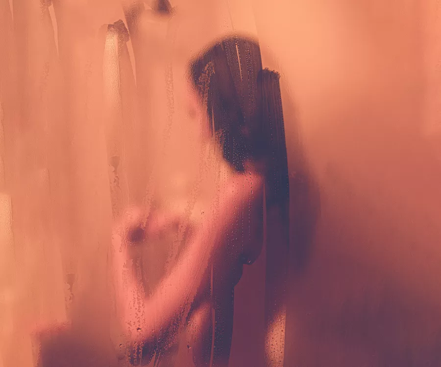 Na pierwszym planie zaparowana szyba ze śladami palców, Przez ślady lekko widoczna kobieta biorąca prysznic