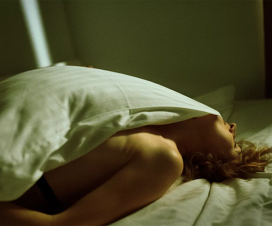 Fotografia kobiety leżącej w białej pościeli, zęby ma zaciśnięte na kołdrze