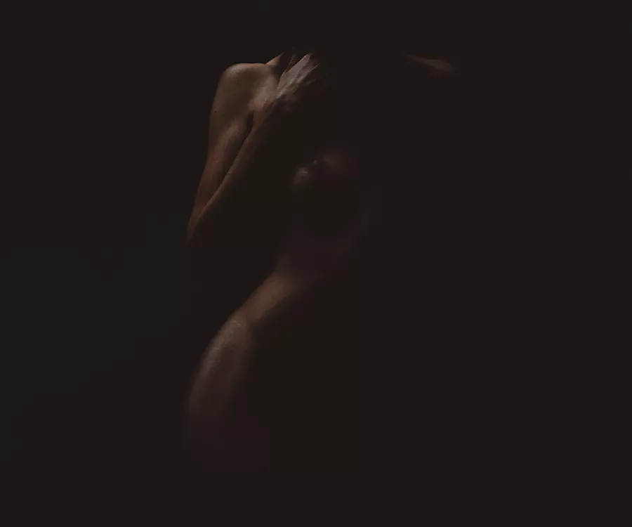 Naga kobieta delikatnie wyłaniająca się z ciemności, widać tylko kawałek biodra, rękę oraz pierś